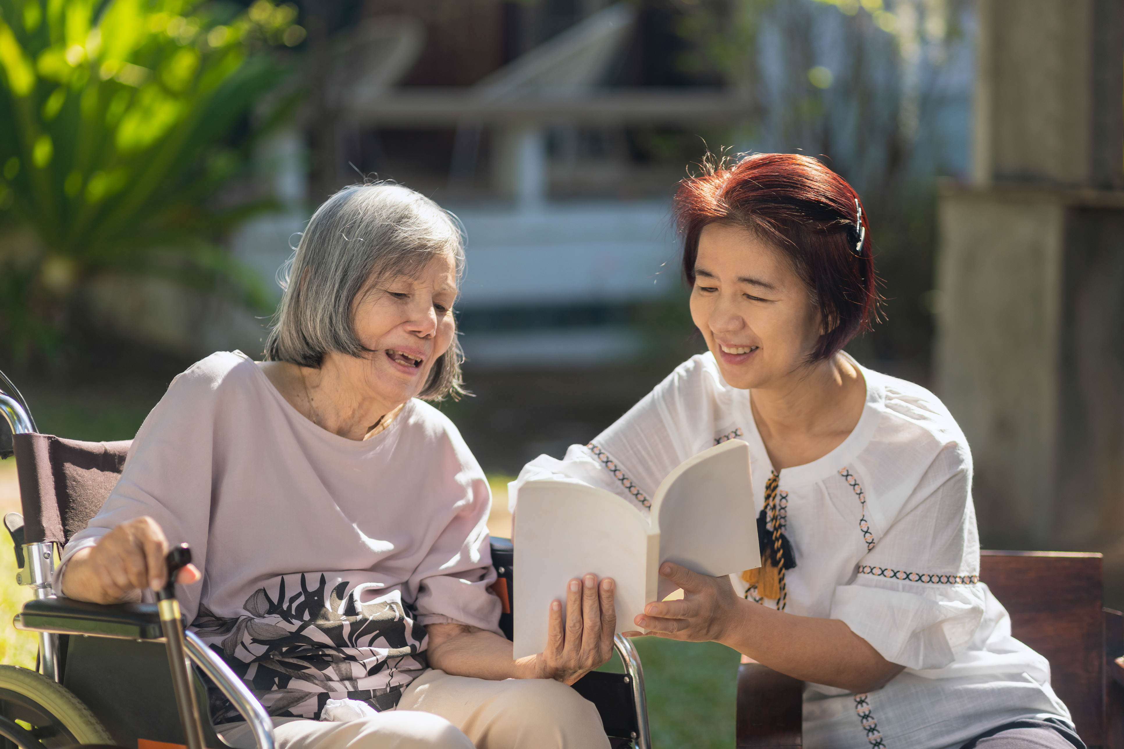 Two Asian women sit outside, the younger woman helps the elder woman read