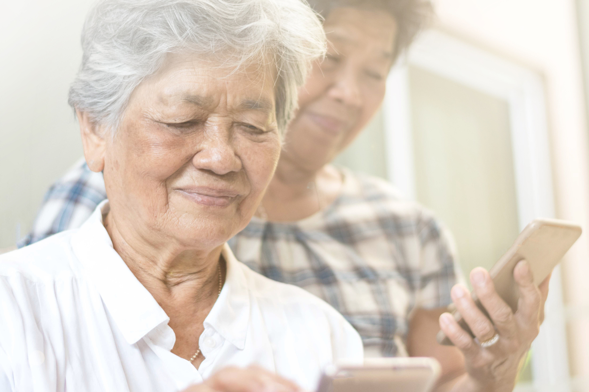 Senior Asian woman with white hair and a white blouse is helped by a caregiver, who supports her from behind. They are both on their phones, trying to read what's on the screen.