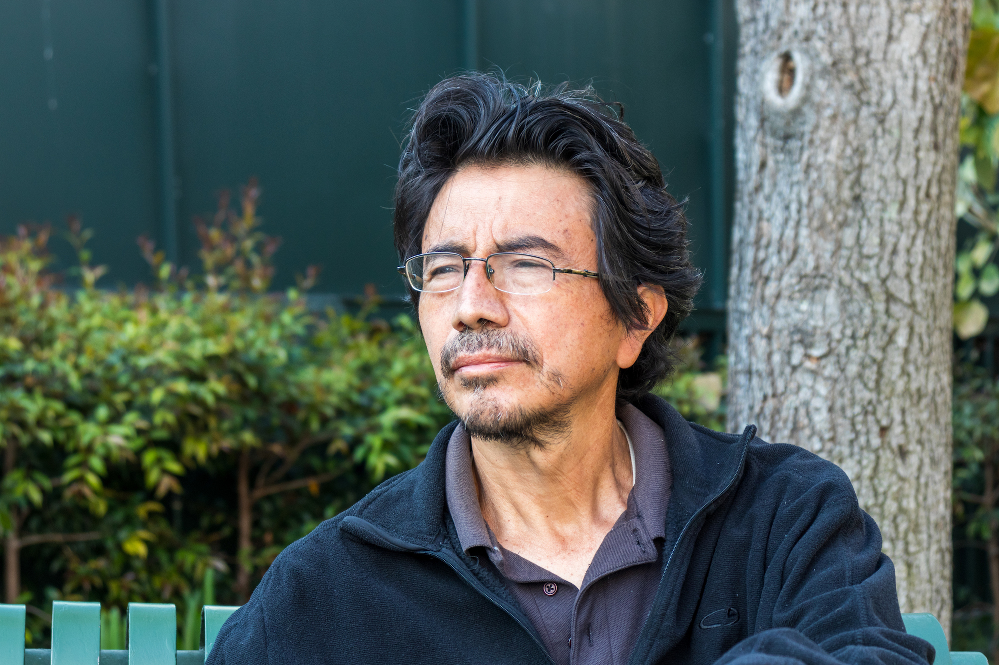 Senior Latino man with glasses and thick dark hair sits outside at a park. He squints, straining to see something in the distance.