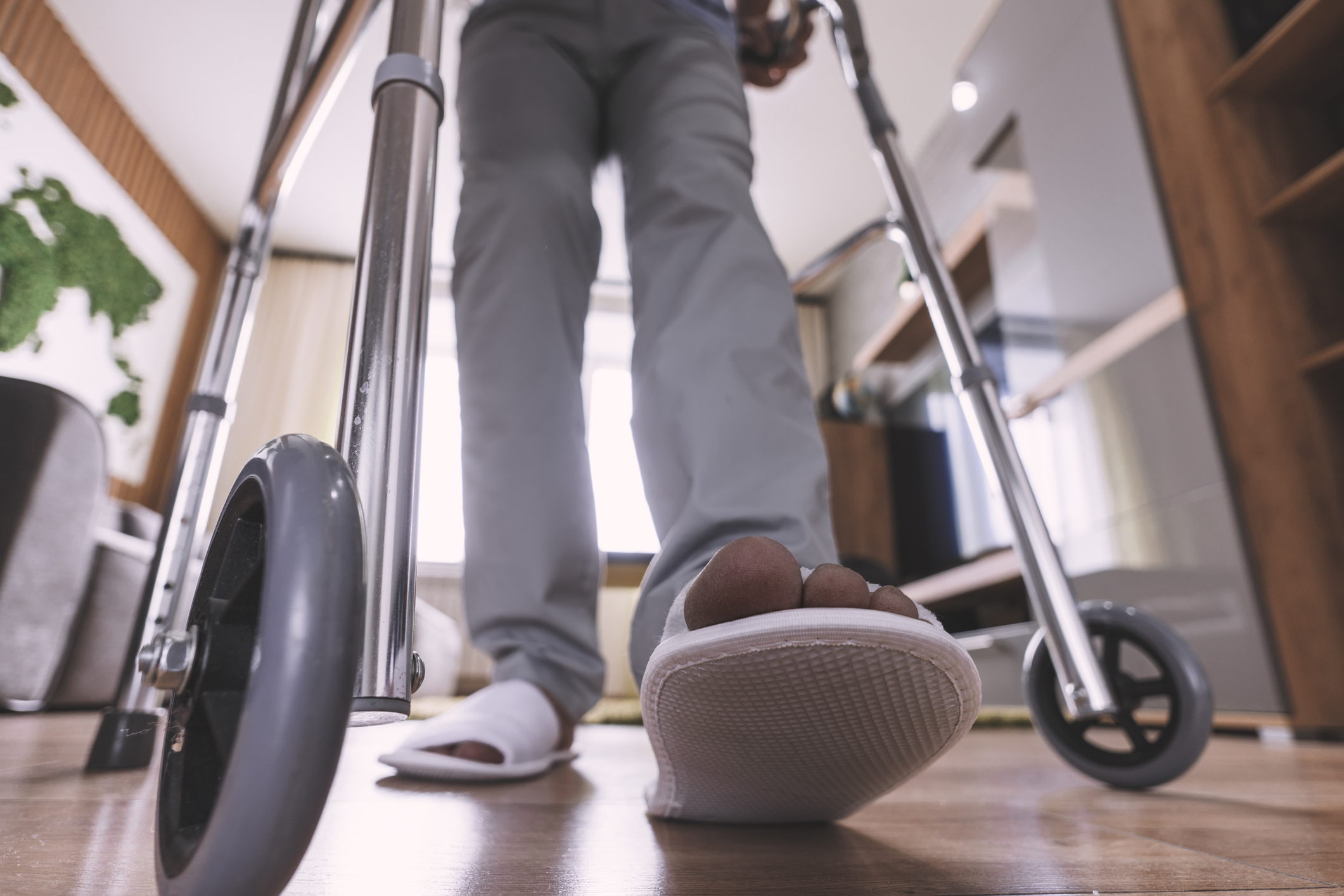 Closeup of white slippers and gray pants as a person uses a walker to navigate around their home