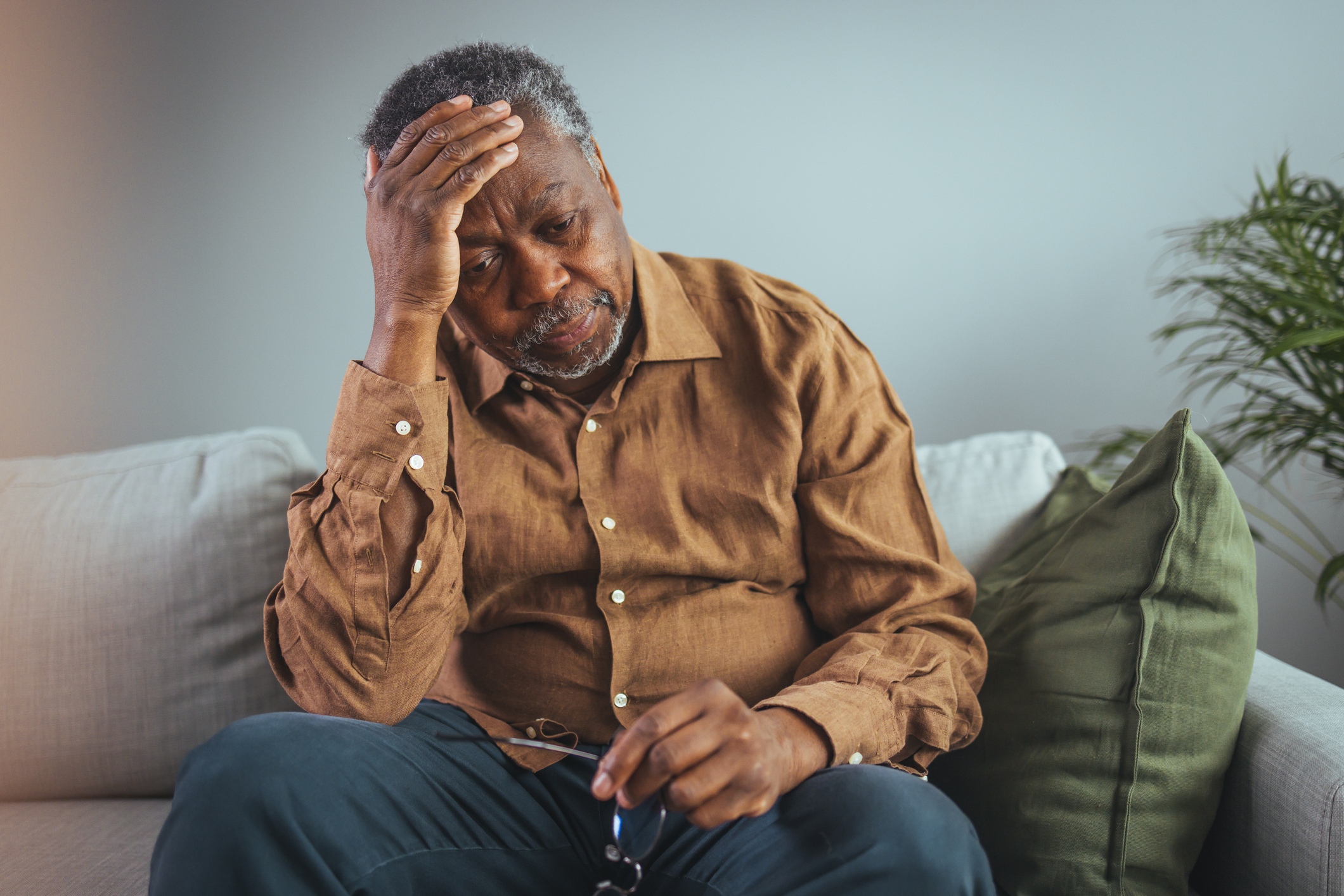 A middle aged Black man in an orange button down shirt sits alone on his couch at home, holding his head in pain in the onset of a stroke