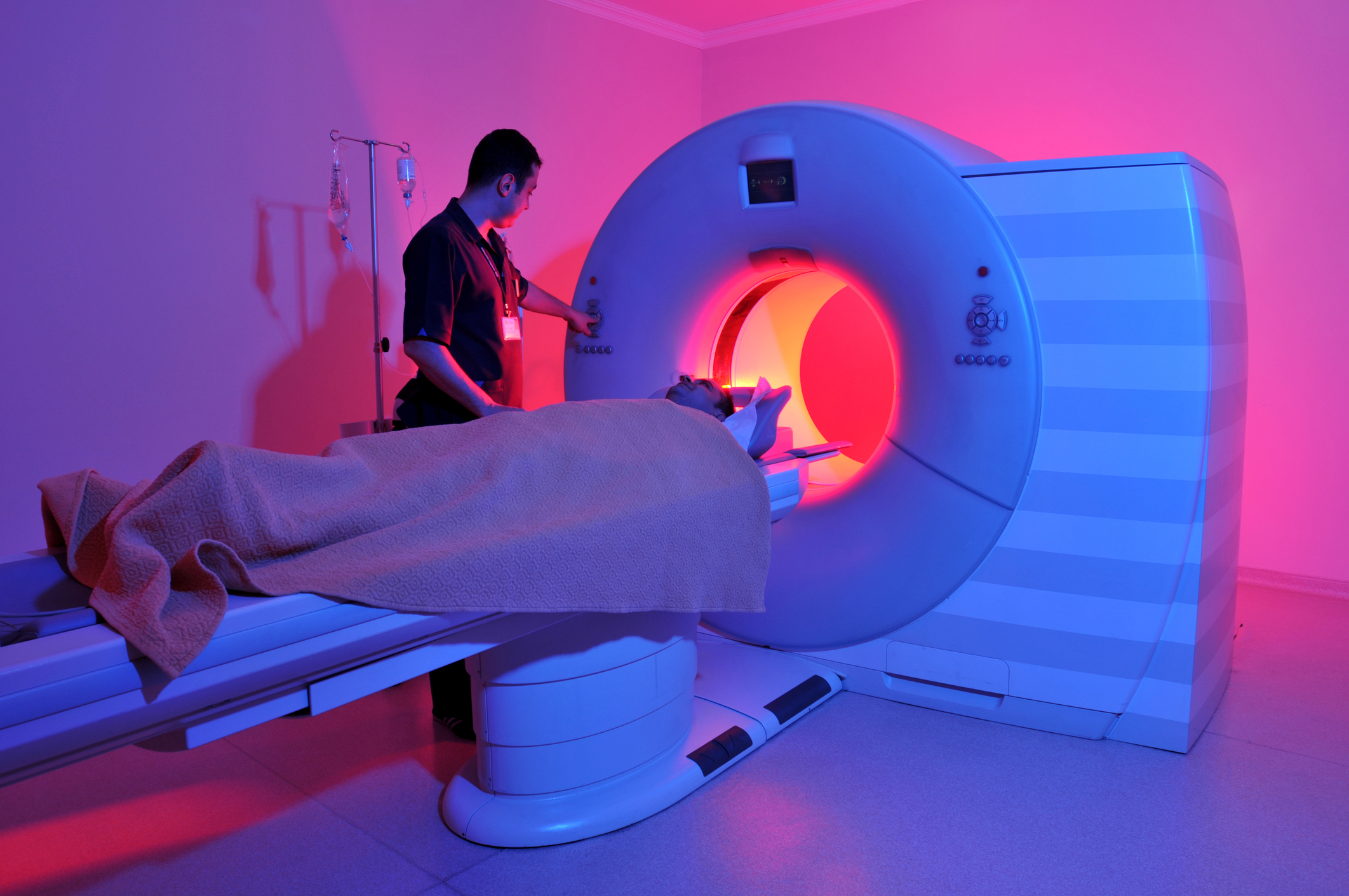A pink-tinted image of a tech in black scrubs starting an MRI scan for a man covered with a thick brown blanket