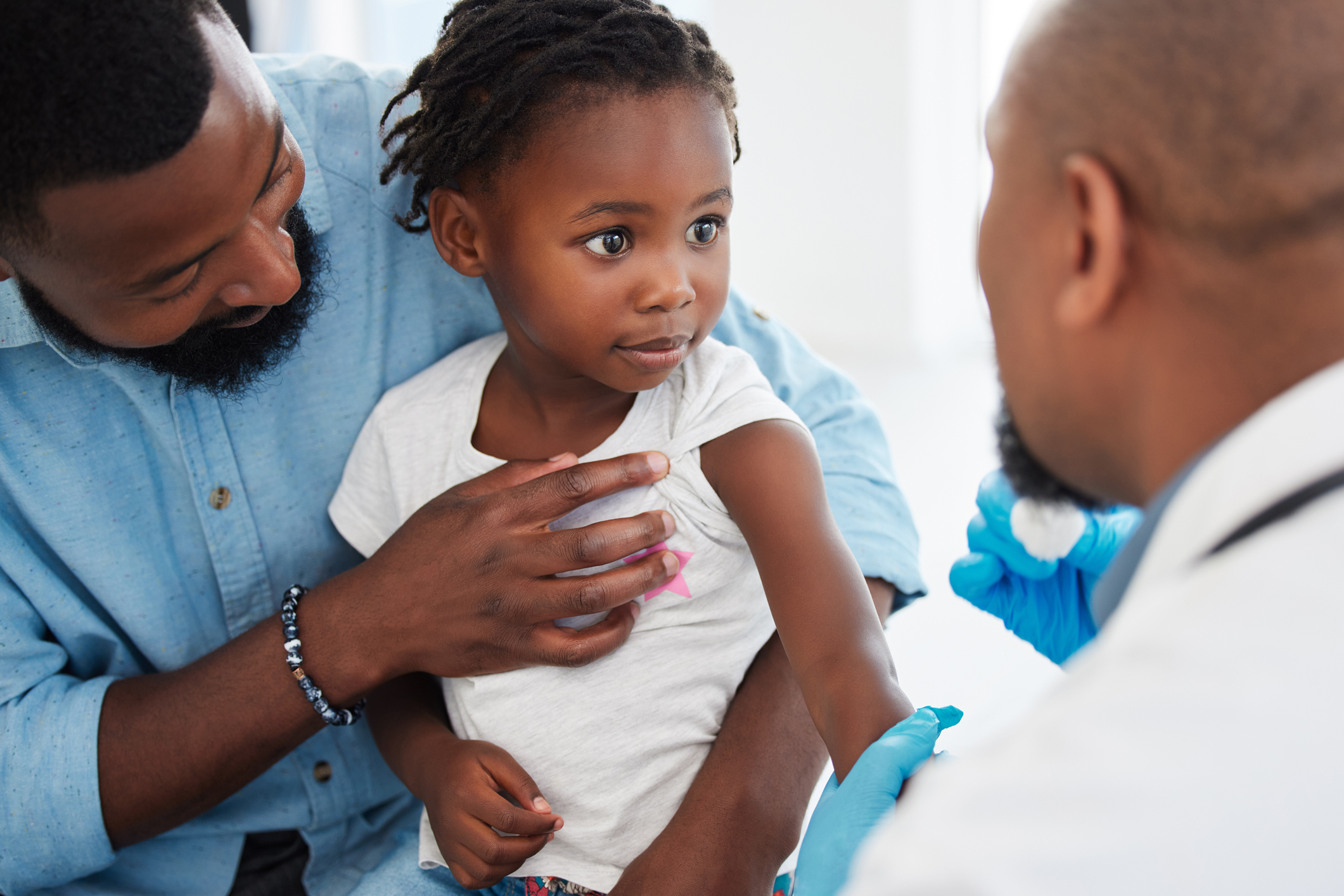 A young Black father protectively holding his toddler daughter while she looks up at a doctor with a nervous expression