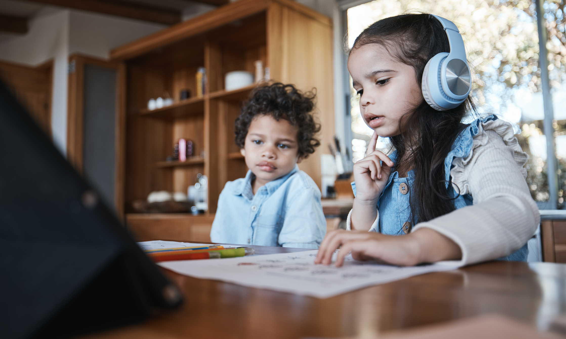 A young girl with wavy brown hair wears noise-cancelling headphones so she can focus on her homework while her younger brother tries to distract her