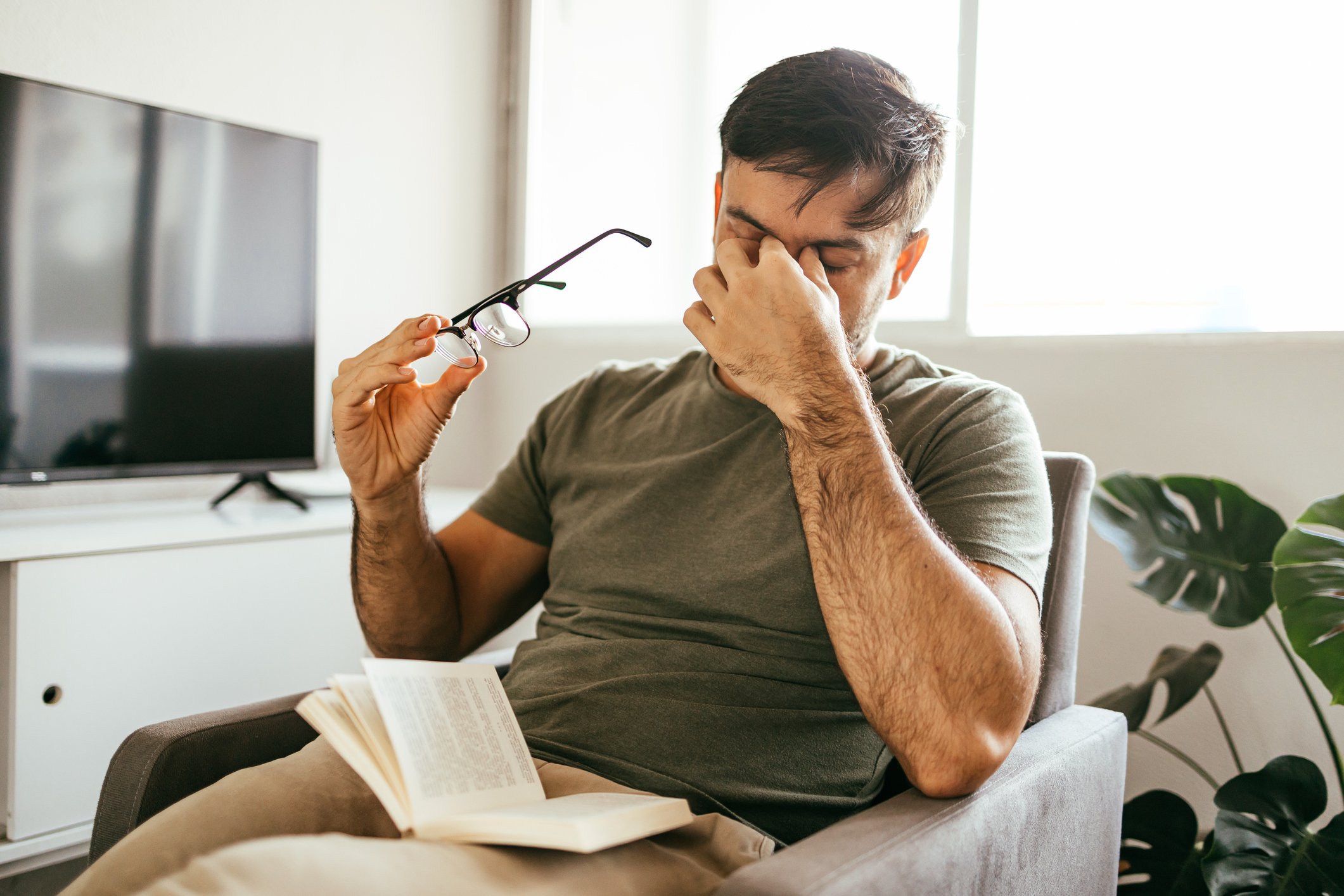 A young man sitting in an armchair at home takes off his glasses to rub his eyes while straining to read a book