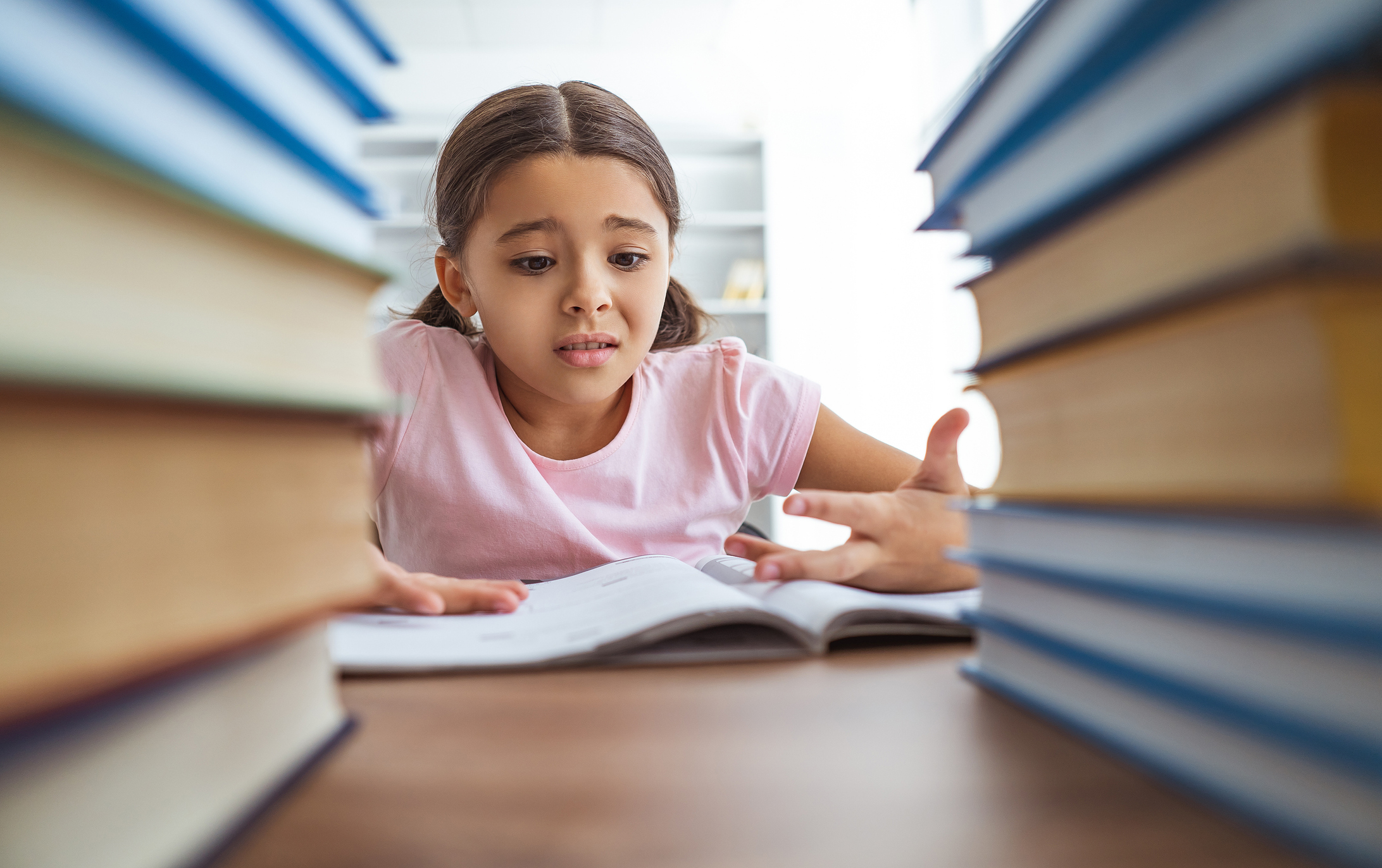 Sitting between two heavy piles of books, an upset girl with light brown hair in pigtails gestures at the worksheet in front of her with frustration and confusion