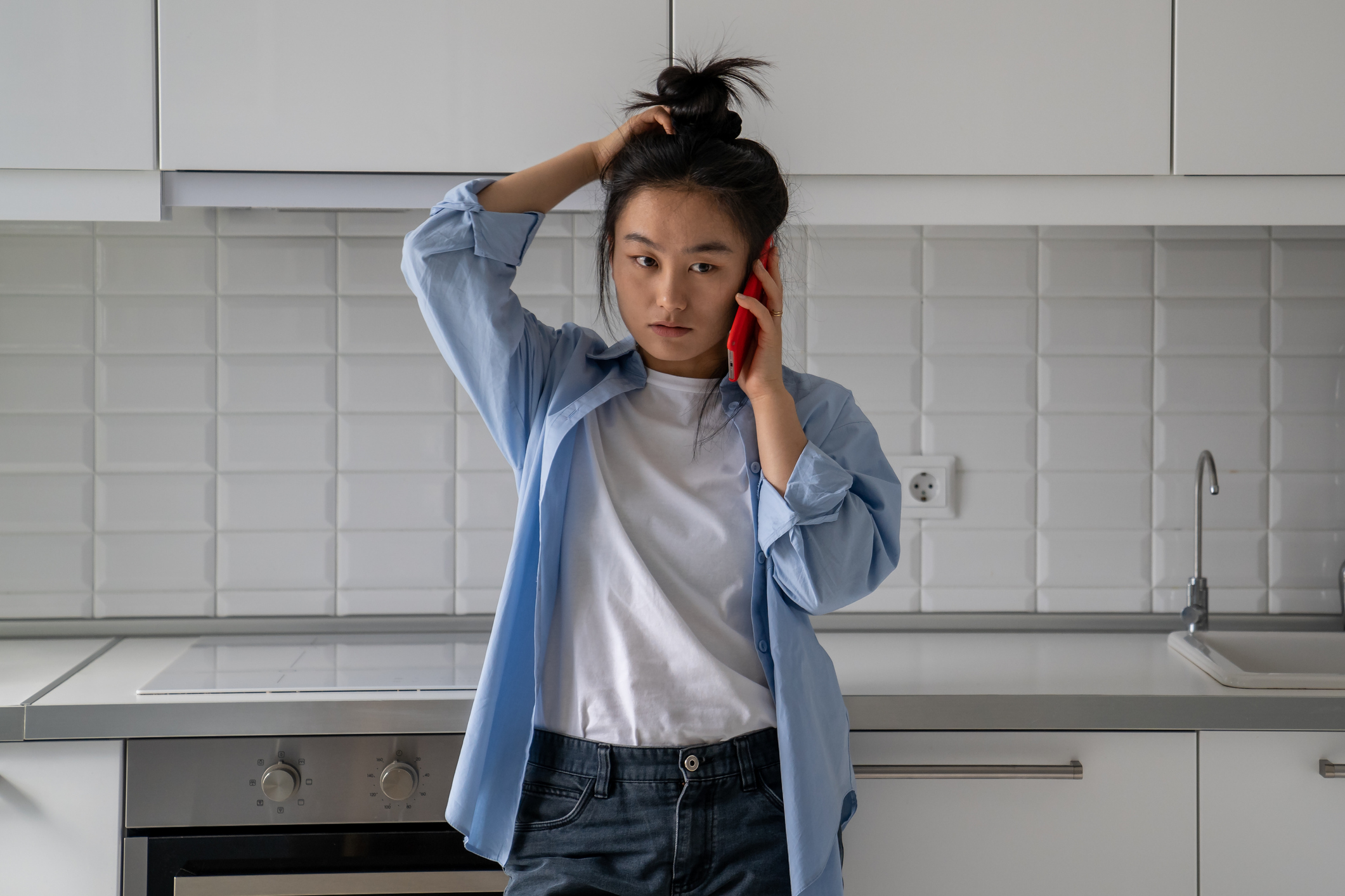 A frustrated young woman rubs her head while standing anxiously in her kitchen, trying to understand what someone says over the phone