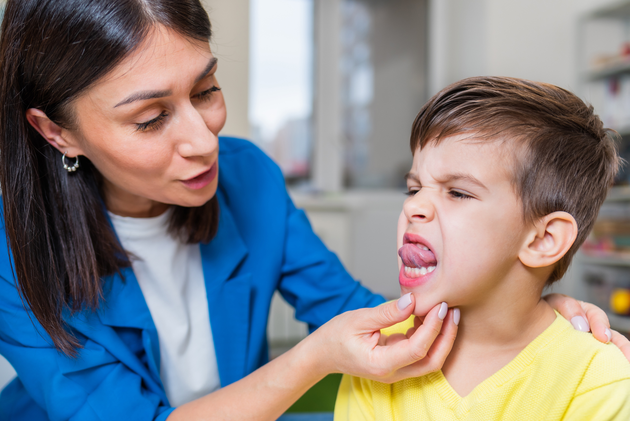 A speech language pathologist in a blue blazer holds a boy's chin to guide him through a muscle-strengthening exercise