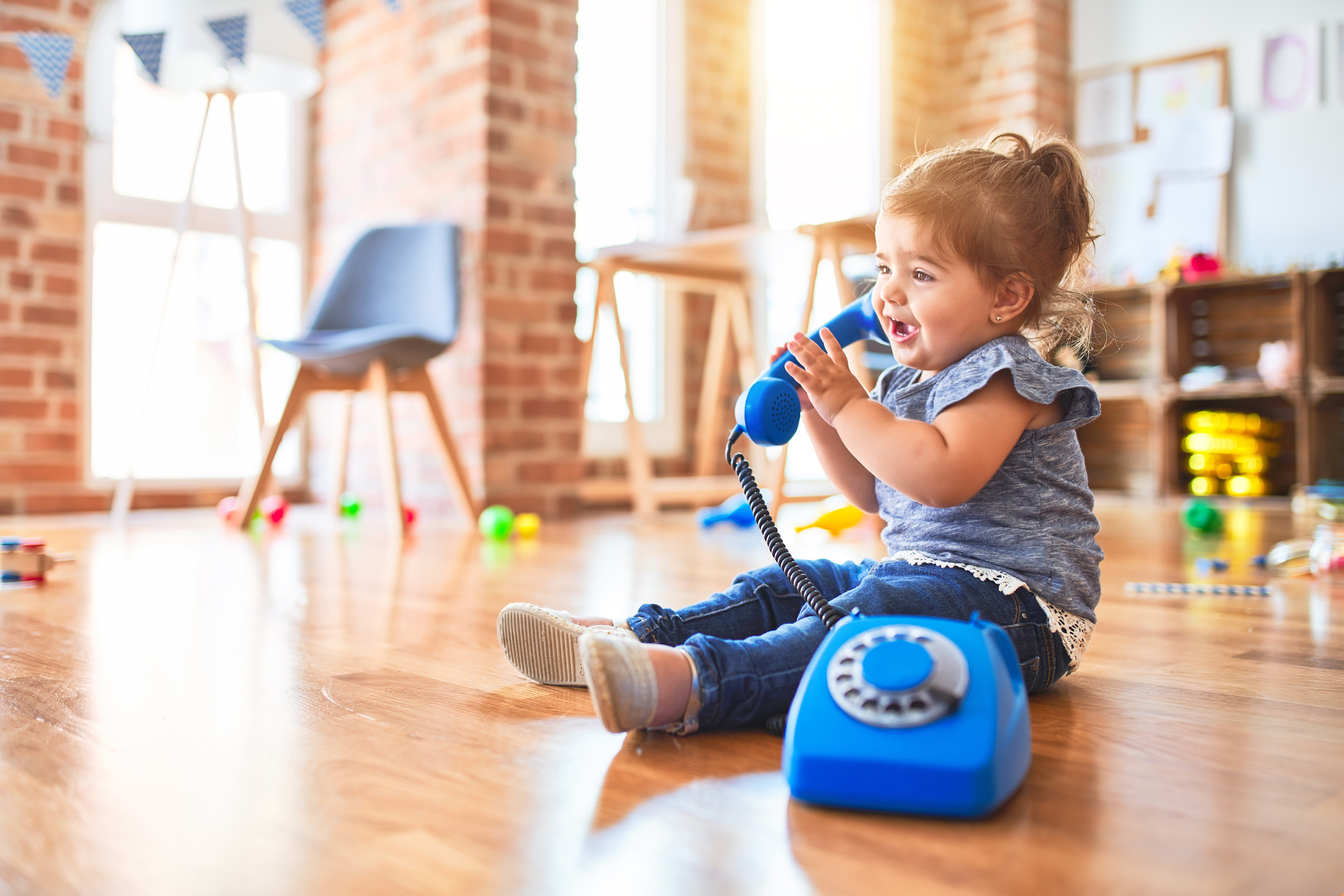 A toddler girl in blue jeans and a frilly blue top plays alone at daycare, pretending to speak into a bright blue toy telephone