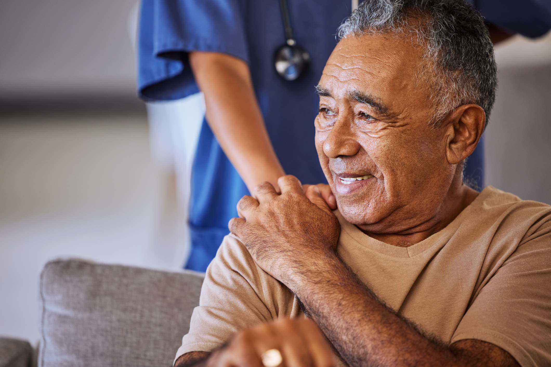 An old man in a nursing home looks off into the distance, confused or concerned, while a nurse in blue scrubs holds his hand