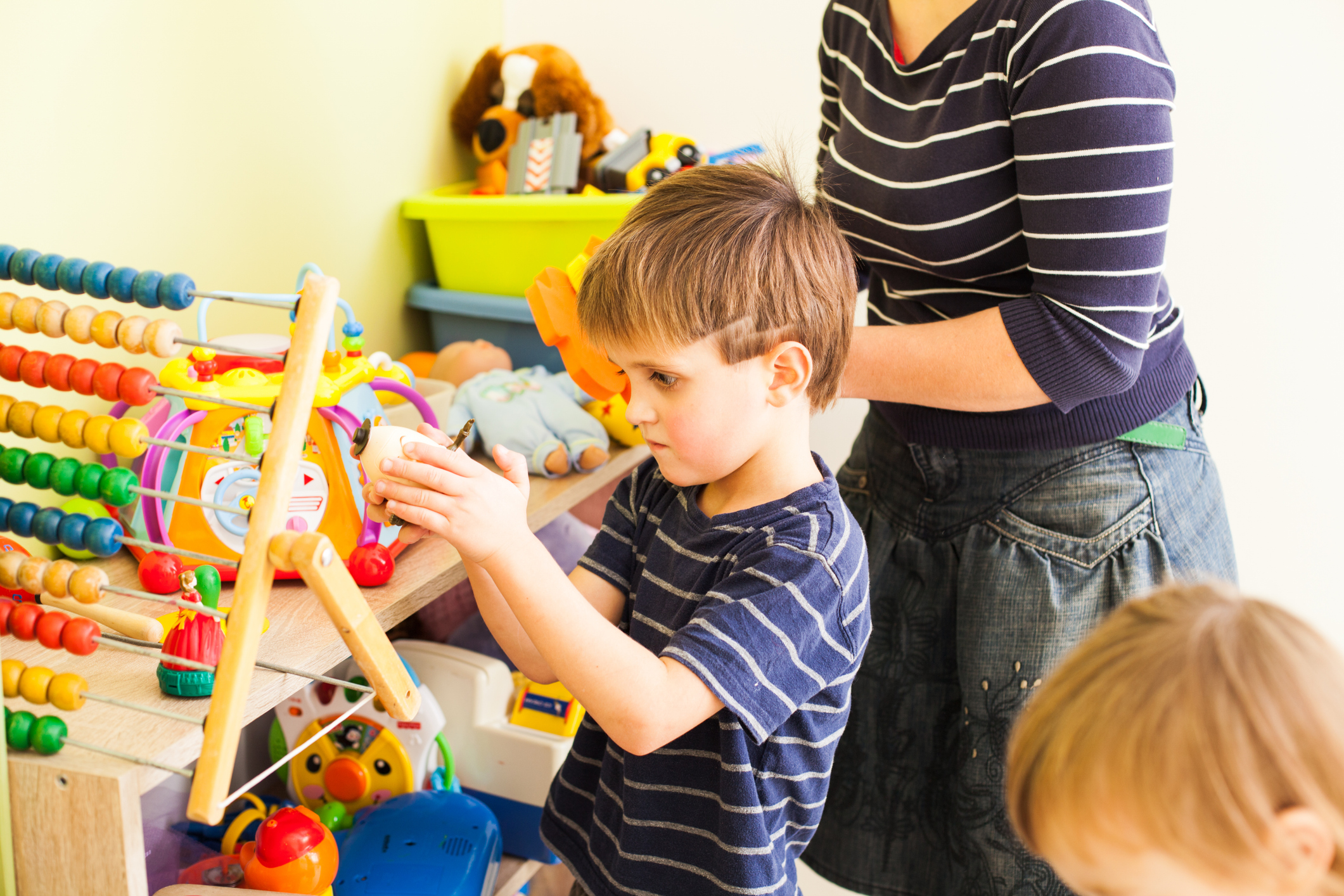 A young white boy with blond hair shaved in a lightning bolt design focuses intently on one toy in front of a shelf full of toys while a white woman in a striped blue shirt and black denim skirt tries to talk to him.