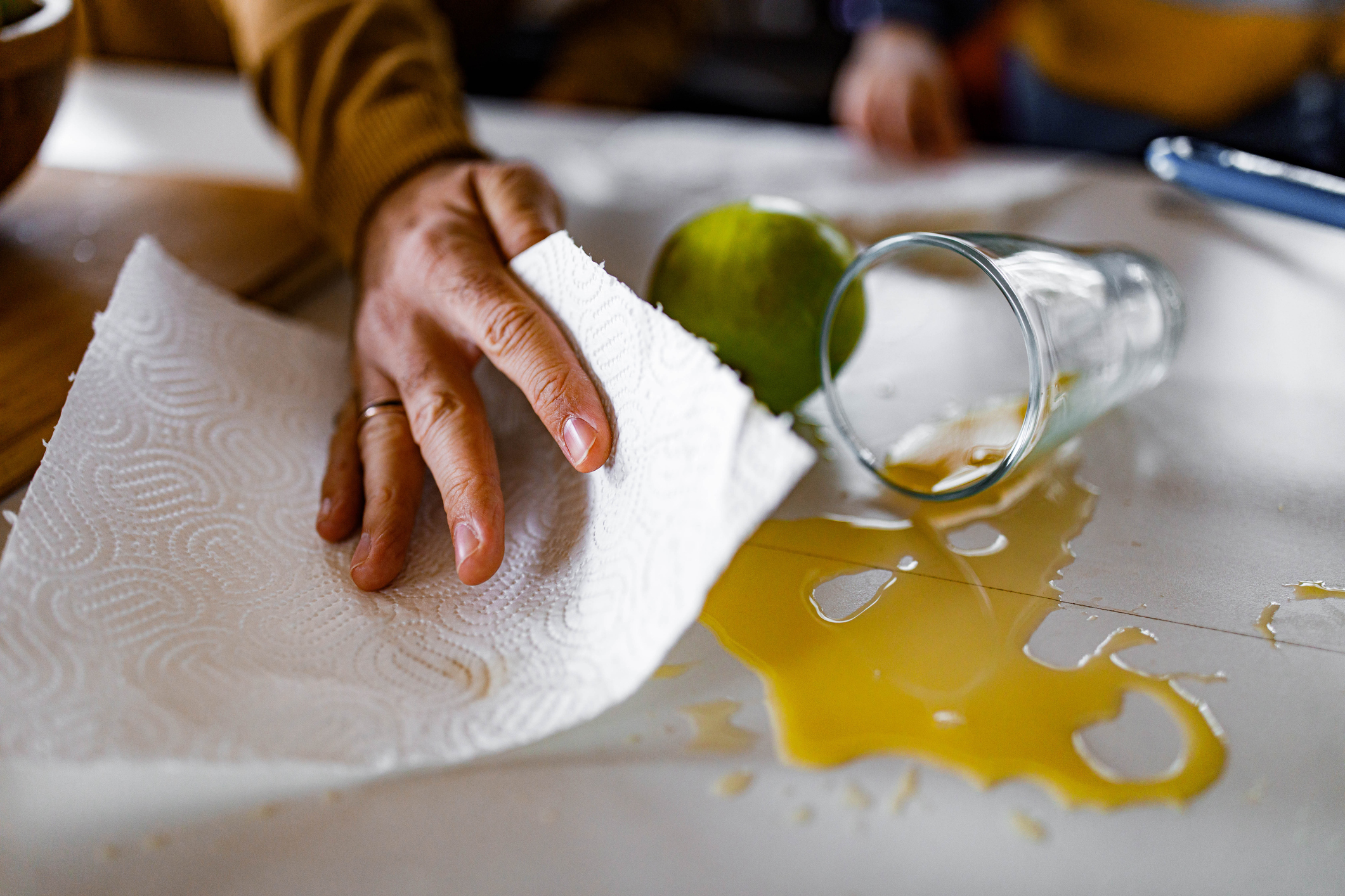 Closeup of a brown-skinned hand as someone uses paper towels to clean up a glass of orange juice spilled on a white table.