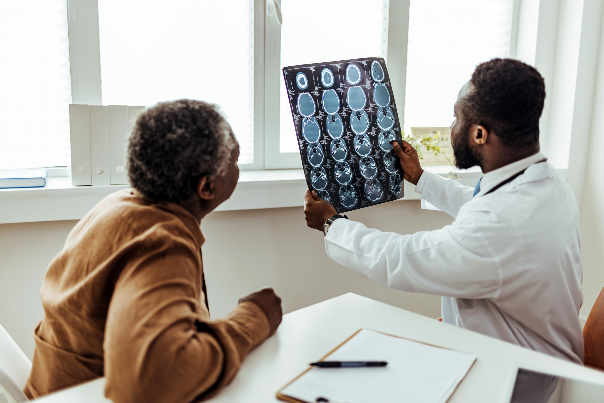 Viewed from behind, a male Black doctor and an older Black man sit in a clean hospital office to review the man's brain scan results
