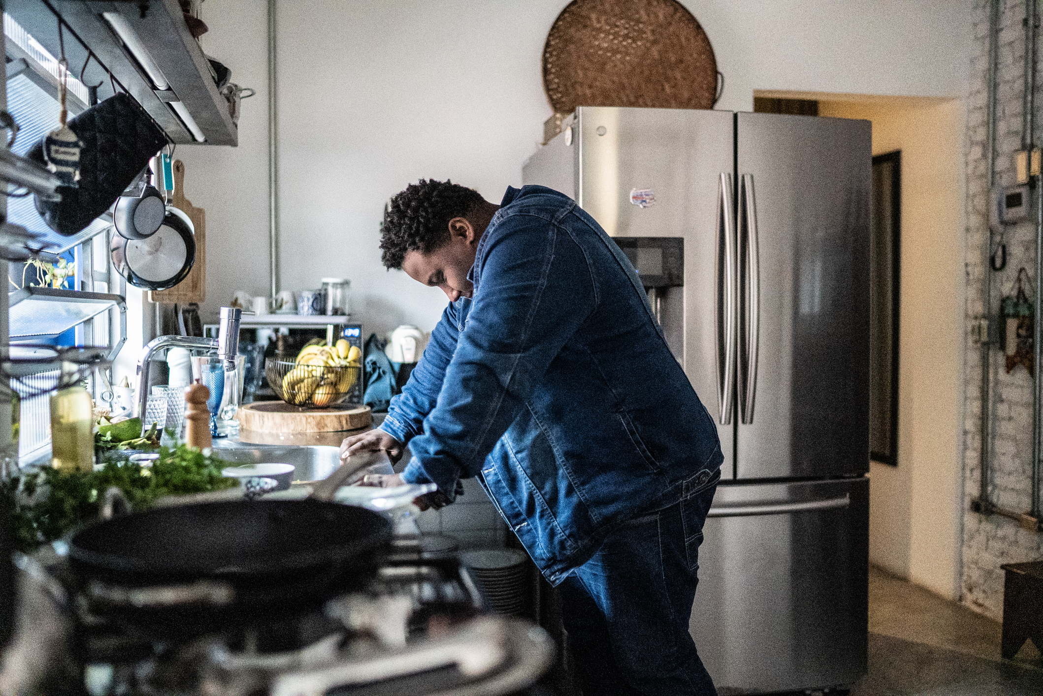A Black man wearing jeans and a denim jacket braces himself against his kitchen counter at home, squeezing his eyes shut while he tries to calm down