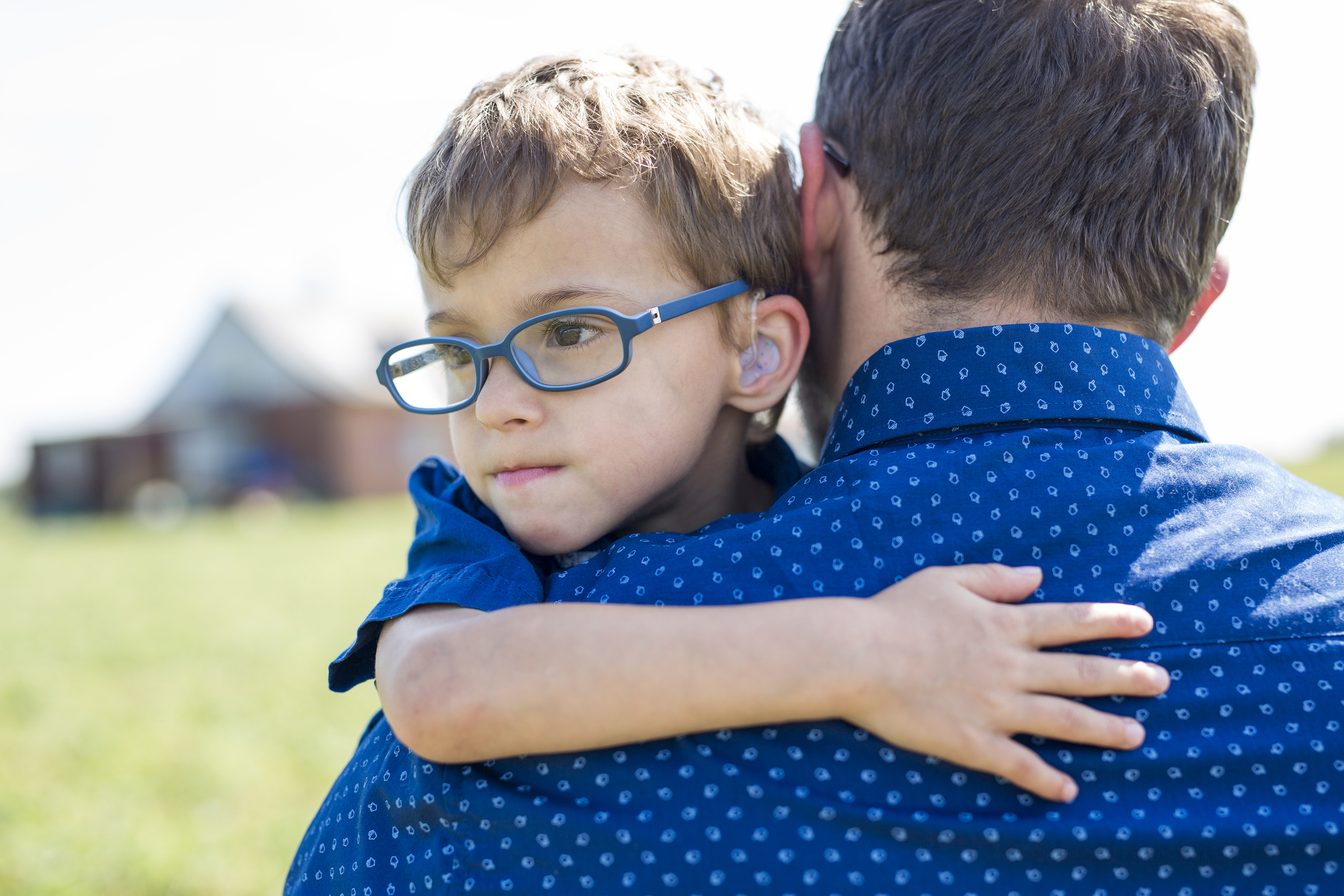 A little boy with pale skin, light brown hair, thick blue glasses, and hearing aids hugs his father, who is carrying him across a field while wearing a bright blue collared shirt