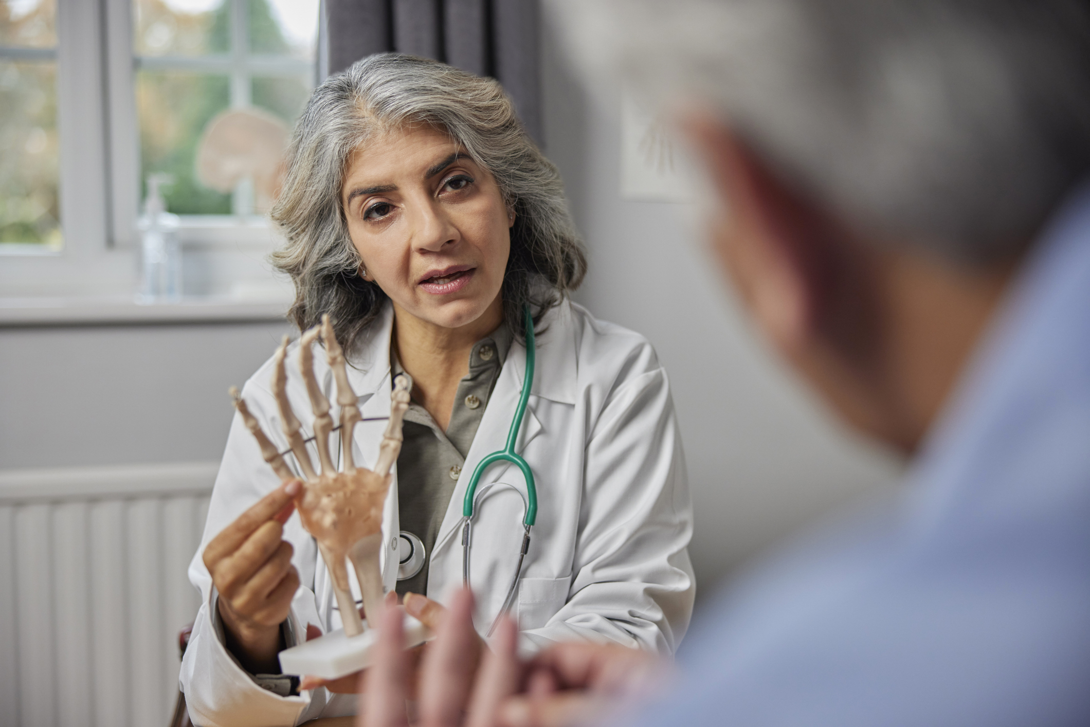 A mature female doctor with brown skin and gray-black hair meets with a blurred-out male patient to explain joint pain using an anatomical model of a hand
