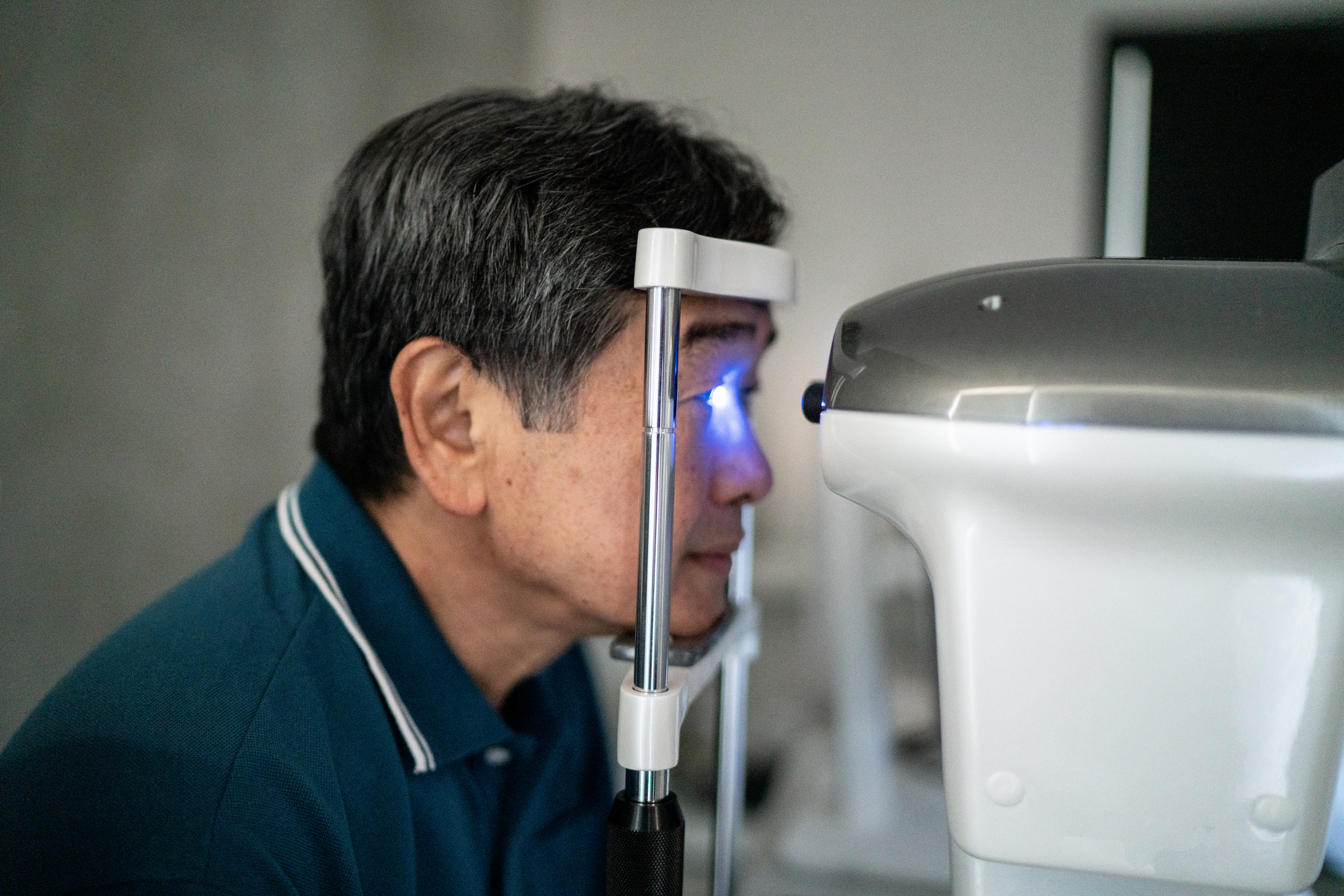A middle aged Asian man in a blue collared shirt leans forward to take an eye test from a machine that shines bright blue light into his eyes
