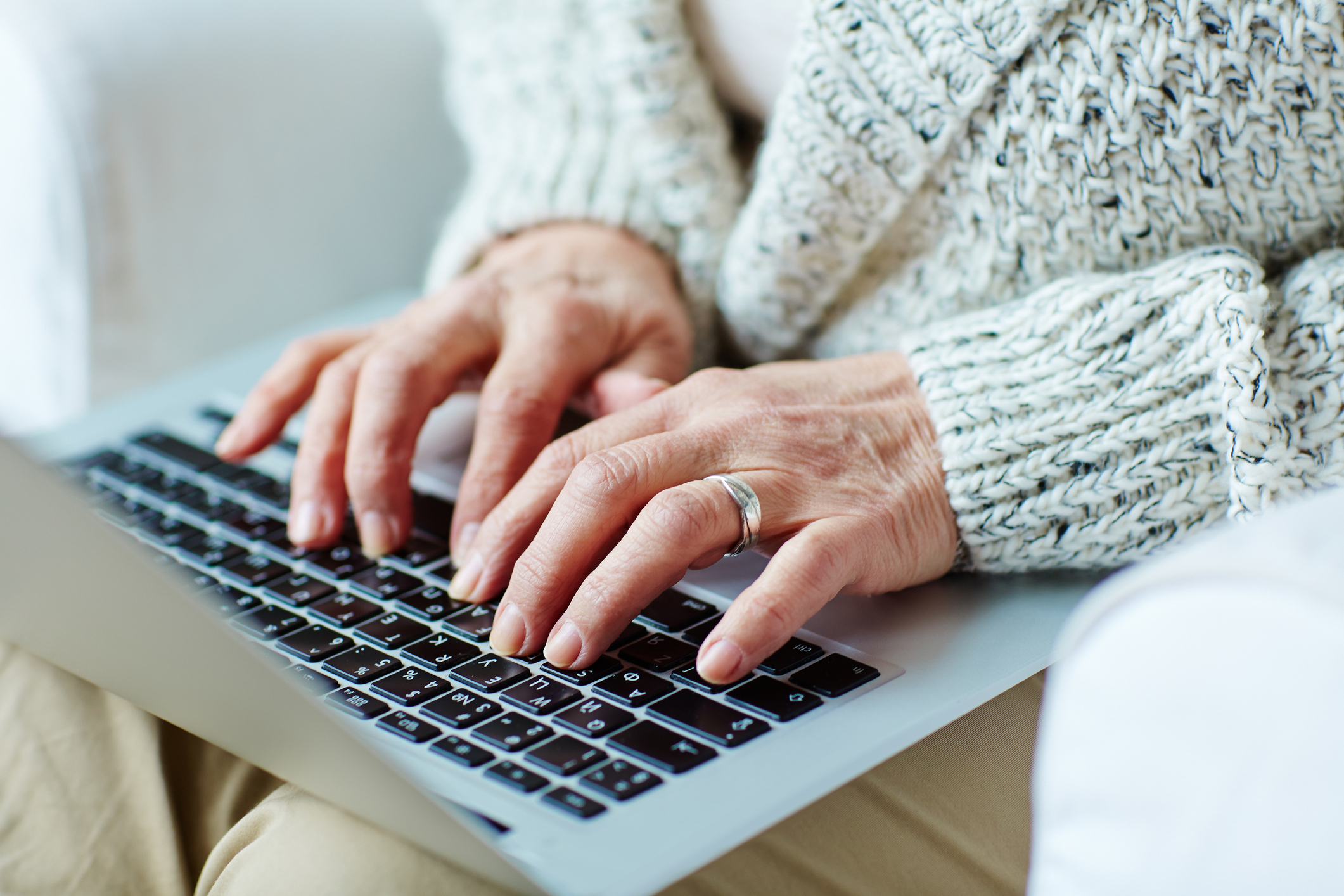 Closeup of an elderly white woman's hands, one wearing a silver ring, as she struggles to type while balancing a laptop in her lap
