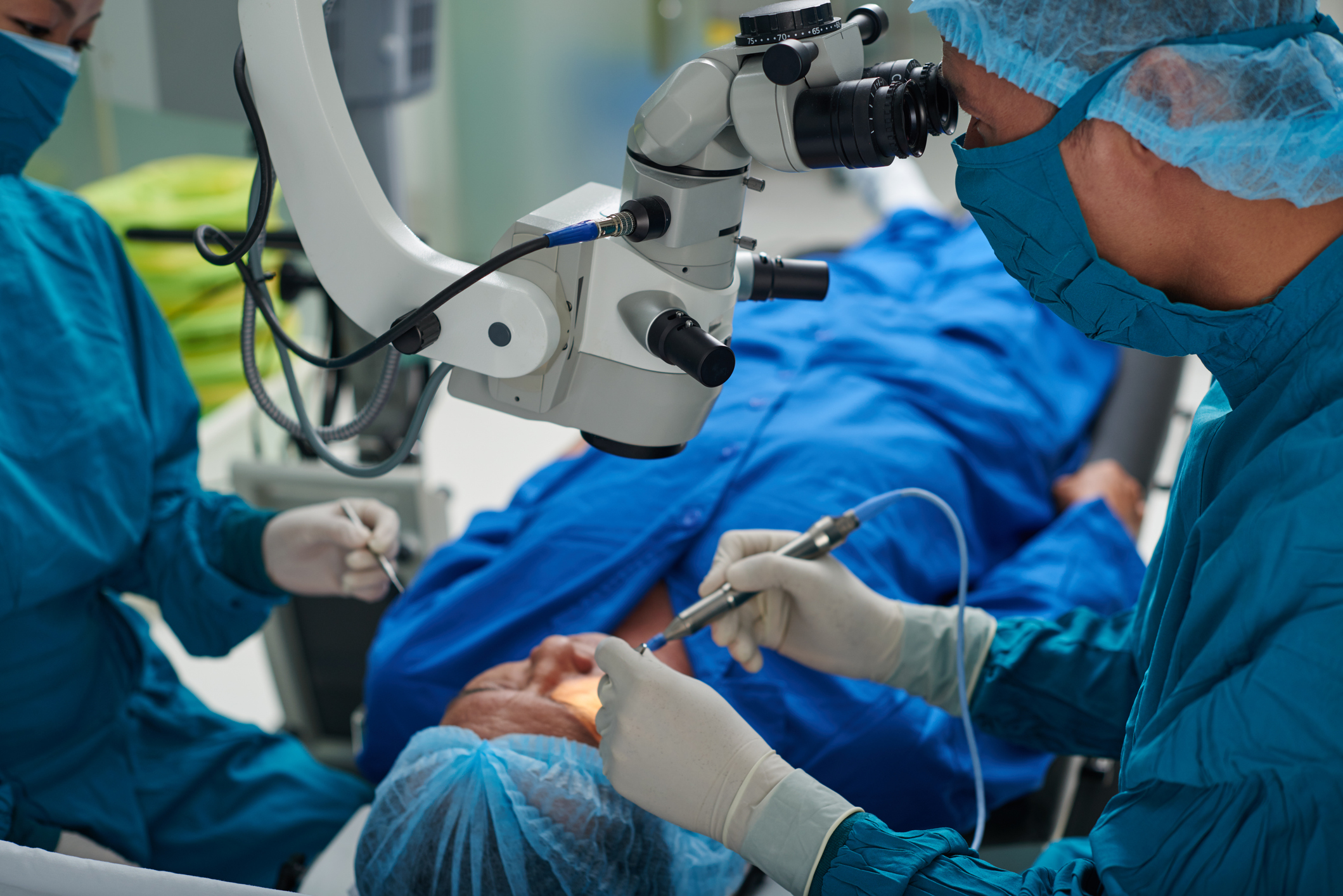 Two doctors, a white man and an Asian woman wearing blue scrubs and surgical masks, perform eye surgery on an out-of-focus patient laying on an operating table.