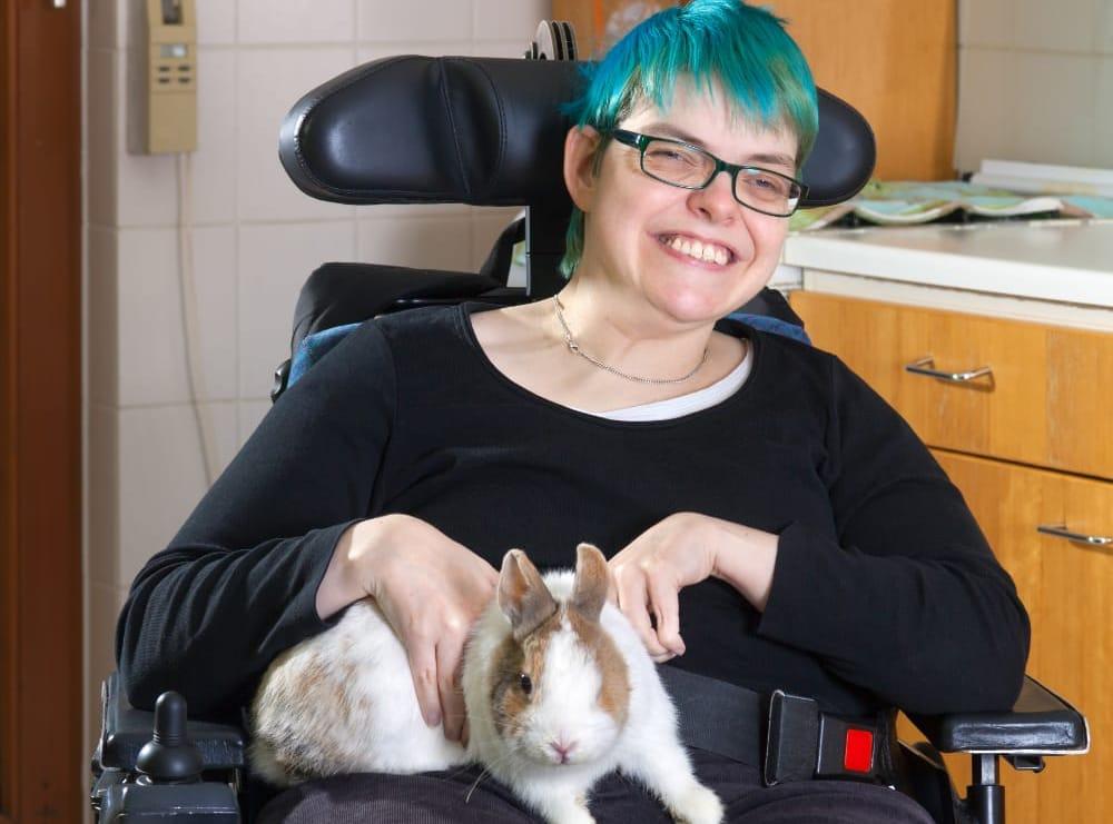White woman with dyed blue hair sits in a wheelchair and smiles as she pets a small rabbit in her lap. 