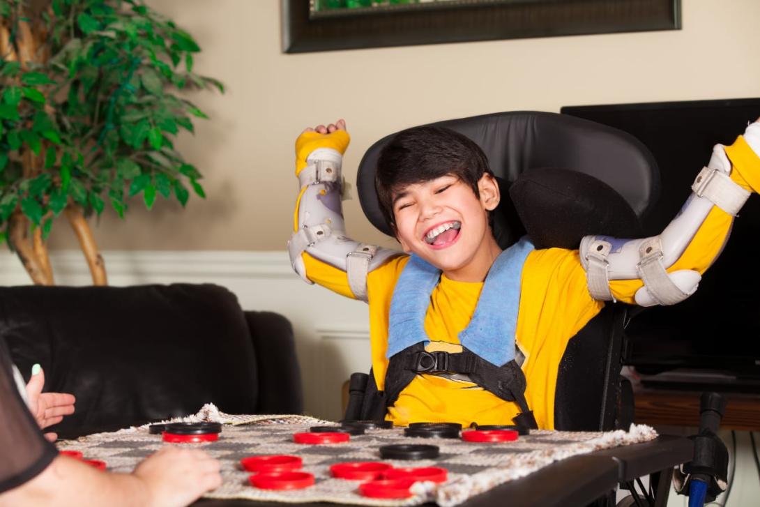 Young Asian boy in a wheelchair and arm braces sits at a table playing a game
