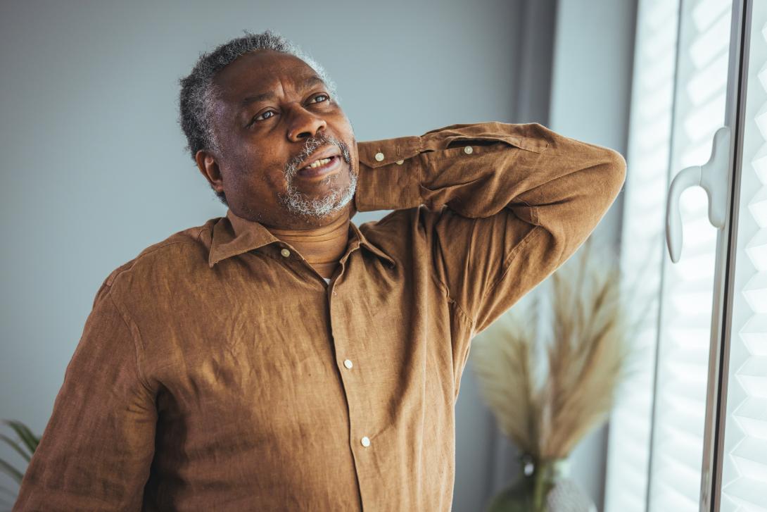 Senior Black man looks out window, rubbing back in pain