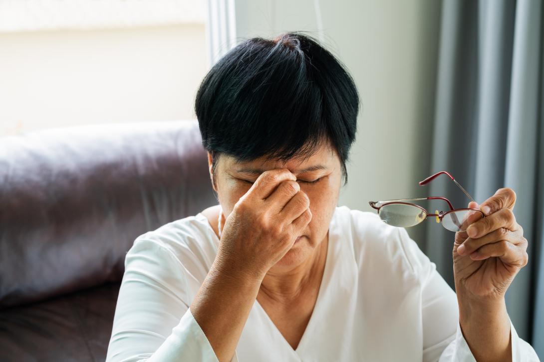 A senior Asian woman sits in her living room on the couch, rubbing her eyes in pain. She has removed her reading glasses and holds them in her hand