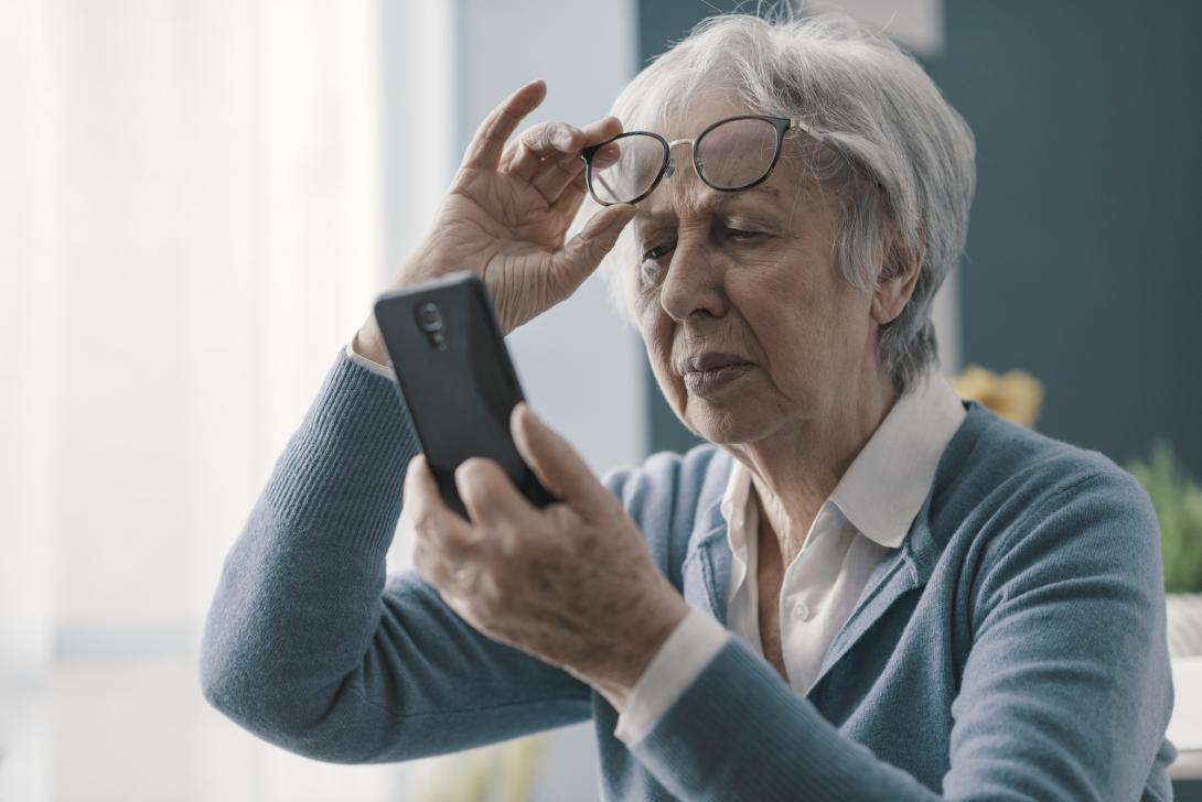 A senior white woman with white hair in a blue sweater sits inside in a dimly lit room. She lifts her glasses and squints at her smartphone, trying to read the screen.