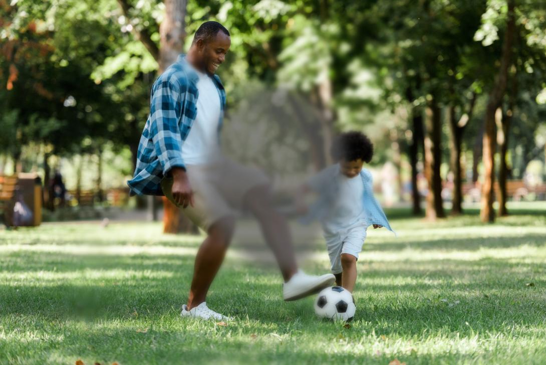 A young Black father and son play with a football outside in a sunny park. There is a dark blurred spot in the middle of the image, to represent the effect of macular degeneration.