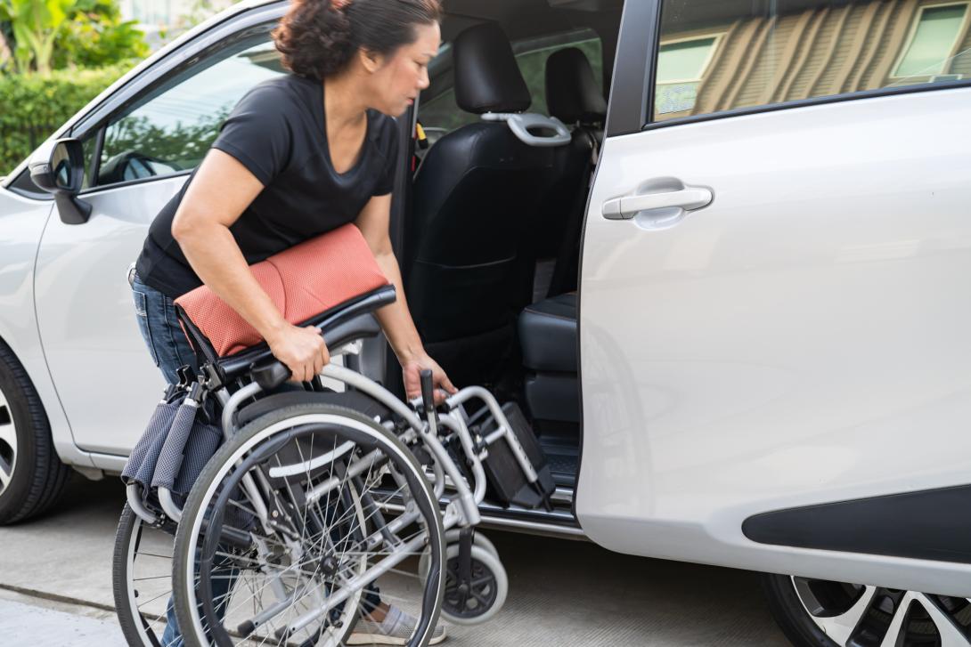  A female caregiver in a black tee shirt and jeans loading a foldable wheelchair into the backseat of an SUV