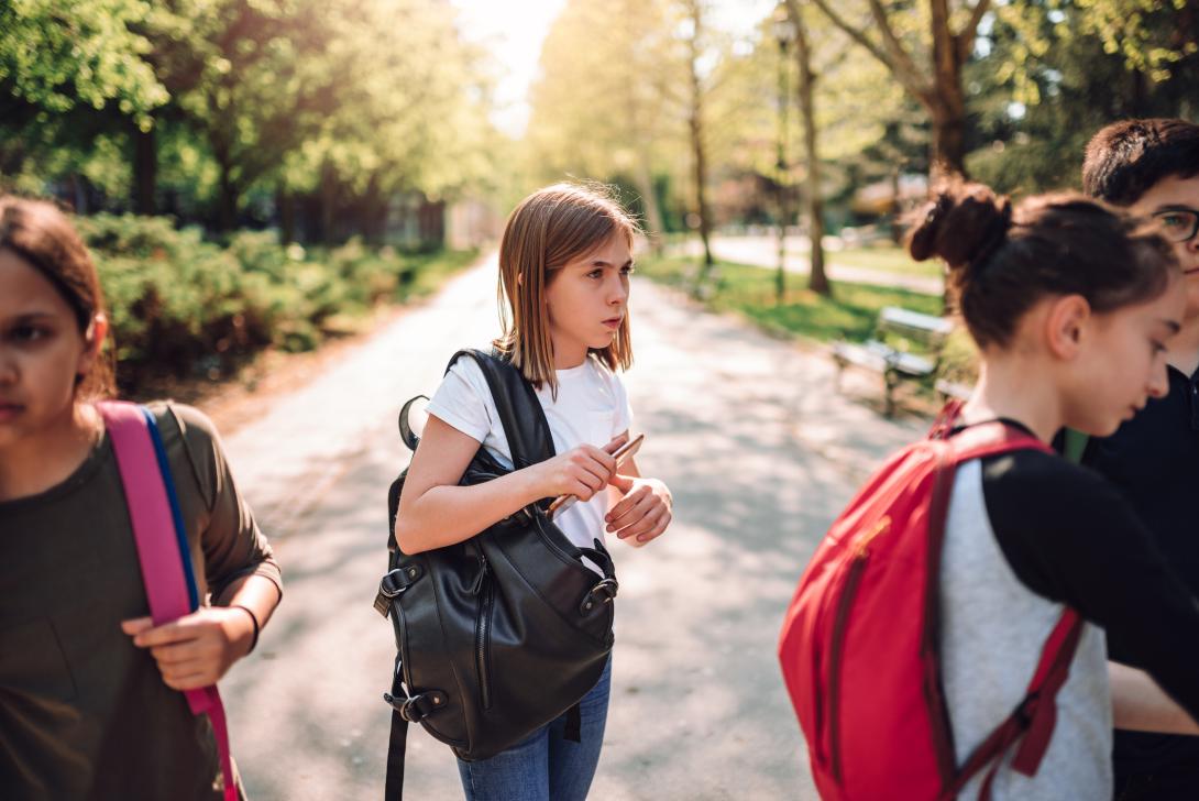 A nervous preteen girl in a white shirt and jeans holding her backpack in the schoolyard, not speaking to other children
